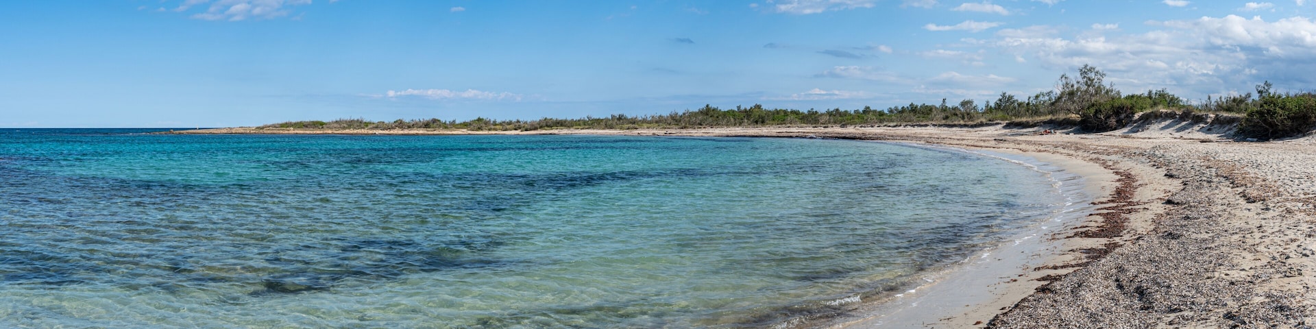 Ein Strand mit klarem türkisblauem Wasser, einem sandigen Ufer und einer fernen Baumlinie unter blauem Himmel mit Wolken.