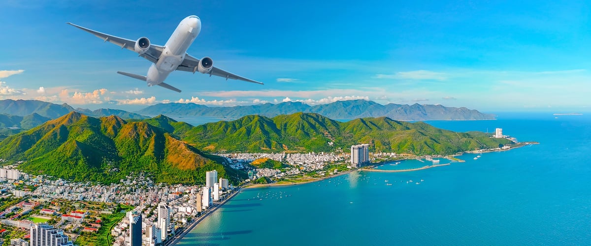 An airplane flying over a coastal city with mountains in the background.