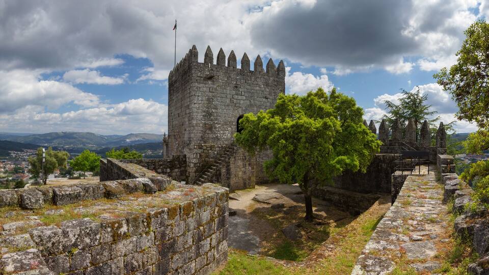 A historic stone castle with a flag, surrounded by lush greenery and a scenic landscape.