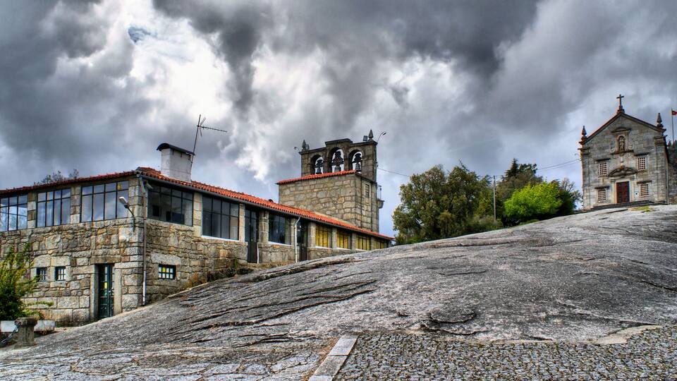 A stone building with a red-tiled roof, a bell tower, and a church with a cross on top.