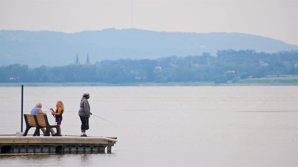 A wooden pier extending into a calm lake with two people fishing. In the background, there is a distant view of a hill with a church spire.