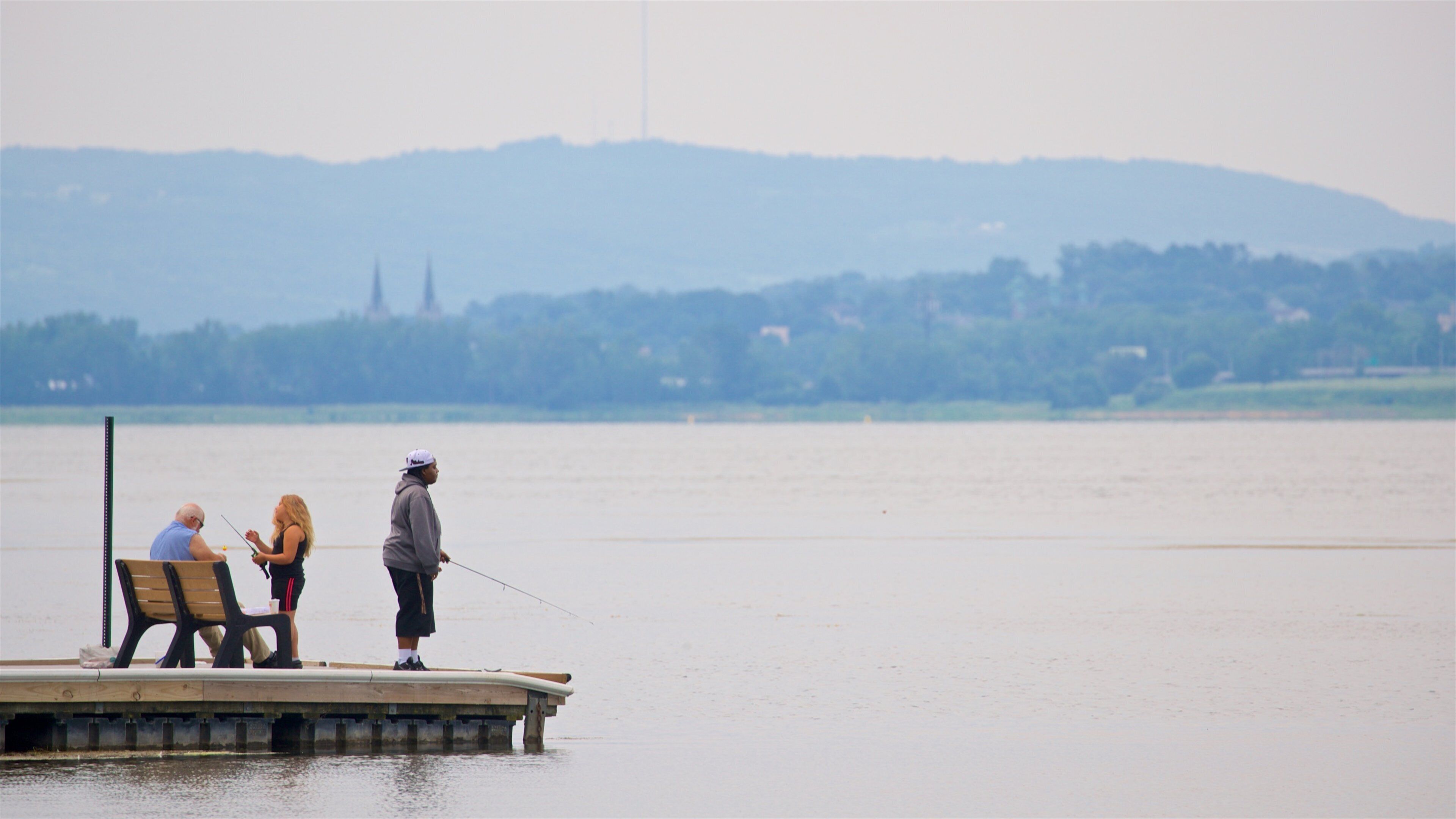 A wooden pier extending into a calm lake with two people fishing. In the background, there is a distant view of a hill with a church spire.