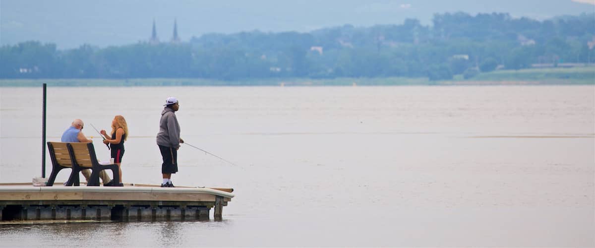 A wooden pier extending into a calm lake with two people fishing. In the background, there is a distant view of a hill with a church spire.