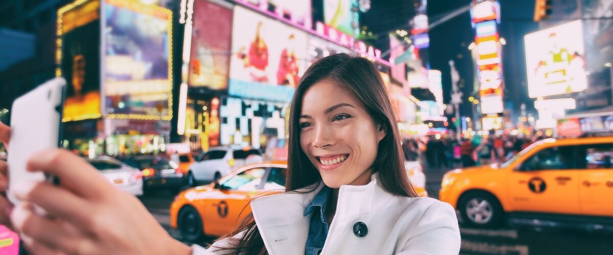 Una persona tomándose un selfie en una zona urbana concurrida, con letreros de neón brillantes y taxis amarillos.