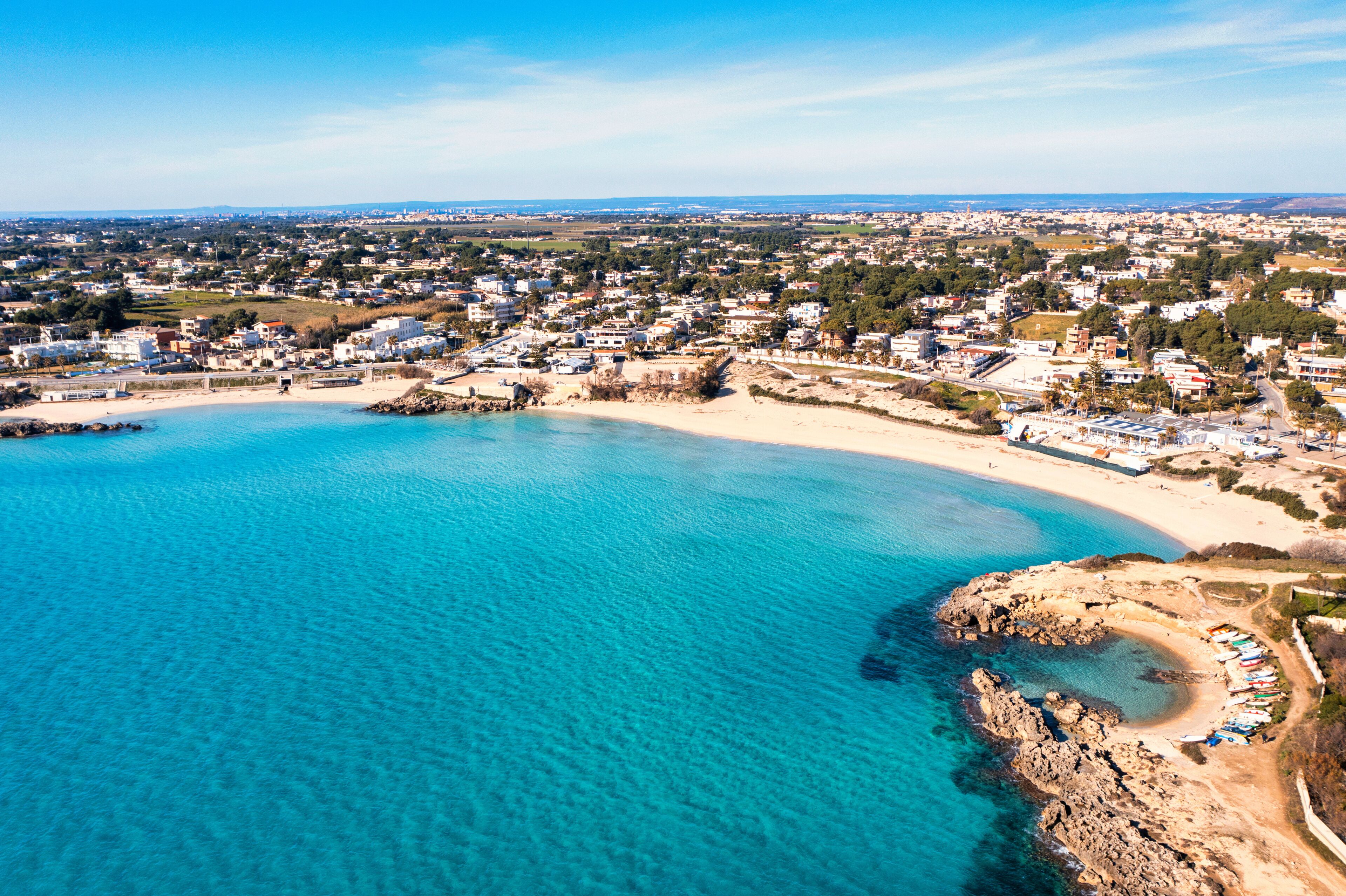 A coastal town with a sandy beach, clear blue waters, and a mix of residential and commercial buildings.