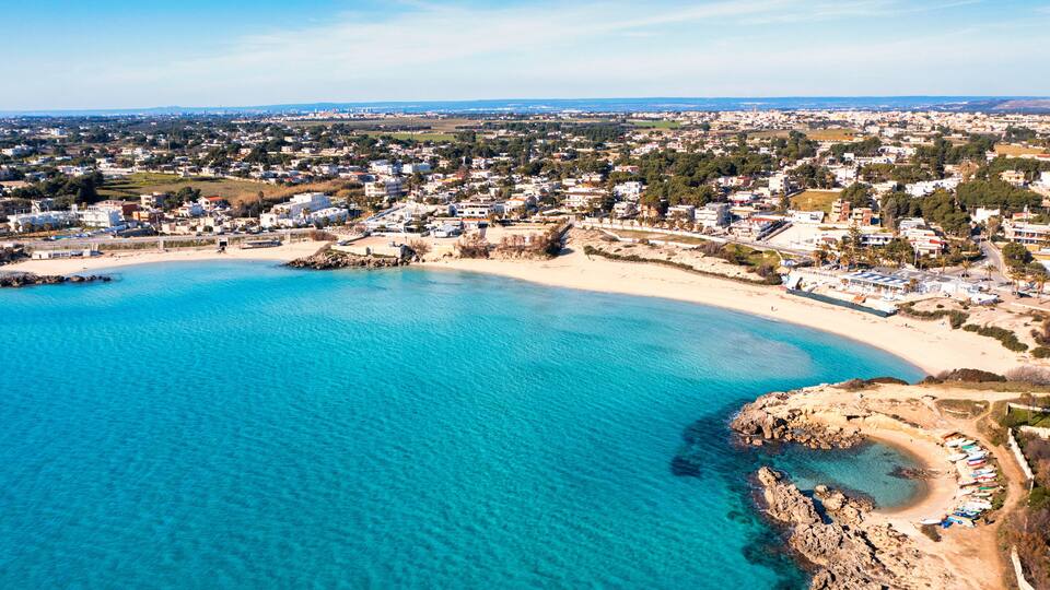 A coastal town with a sandy beach, clear blue waters, and a mix of residential and commercial buildings.
