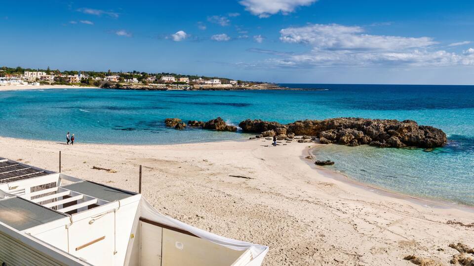 A beach with clear turquoise waters, rocky outcrops, and a white building in the foreground.
