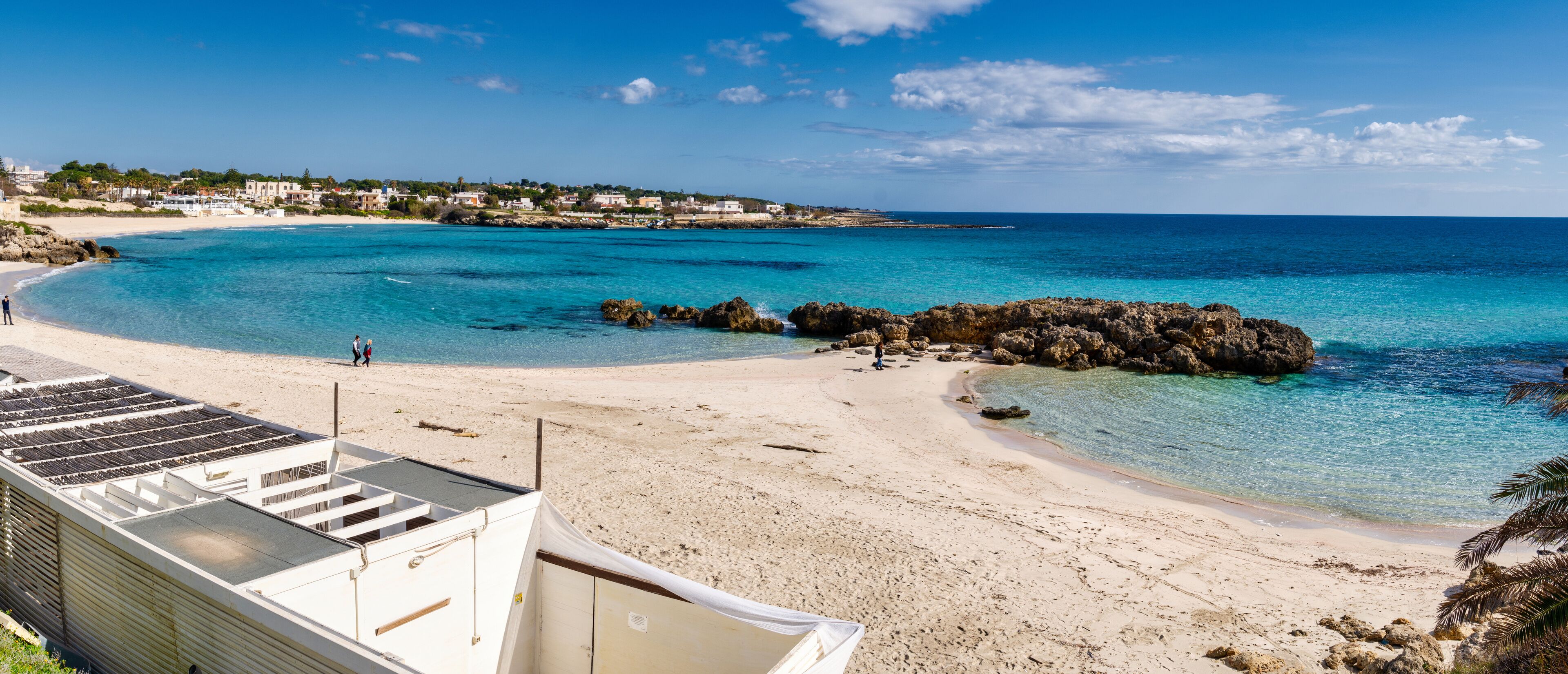 A beach with clear turquoise waters, rocky outcrops, and a white building in the foreground.