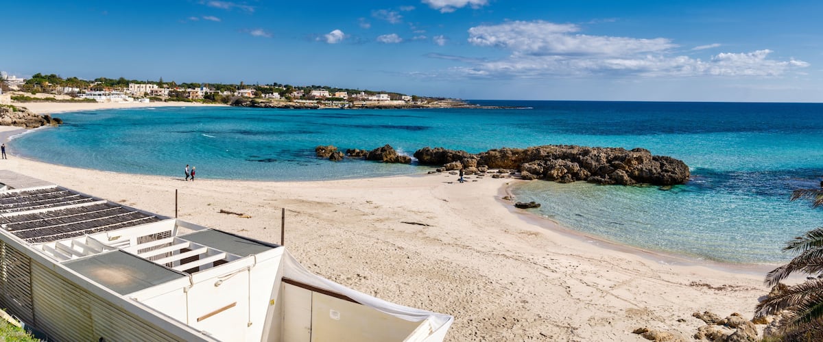 A beach with clear turquoise waters, rocky outcrops, and a white building in the foreground.