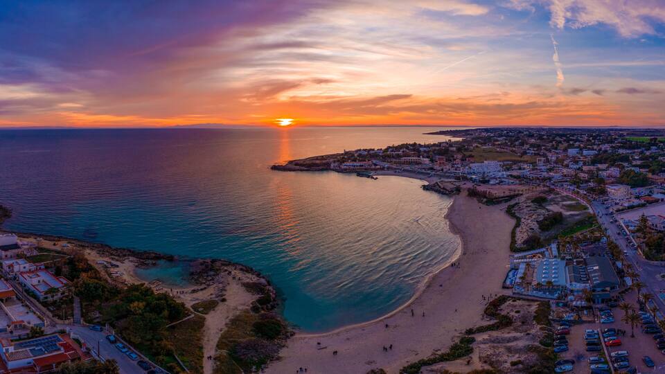 A coastal town at sunset with a beach, a pier, and a clear sky.