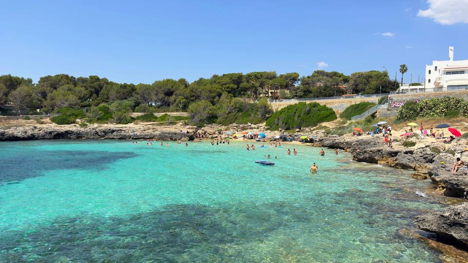 A coastal beach with clear turquoise water, rocky shore, and people swimming and sunbathing.