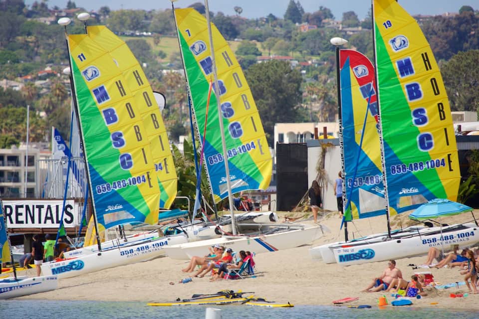 Uma praia com veleiros, pessoas relaxando e um letreiro que diz 'Boat Rentals'.
