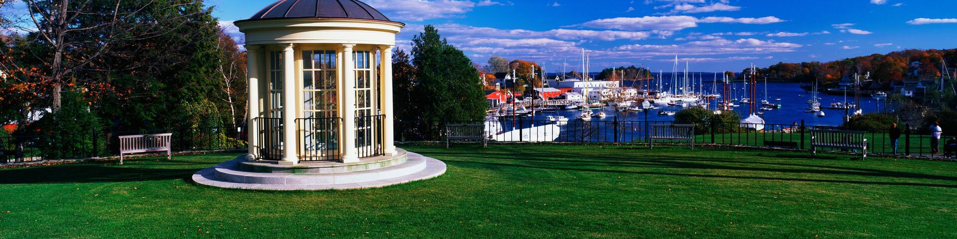 This is the gazebo of the public library. It has a view of the harbor with autumn foliage surrounding it. It is set on a green lawn and the gazebo has off-white columns, a black wrought iron railing and windows.