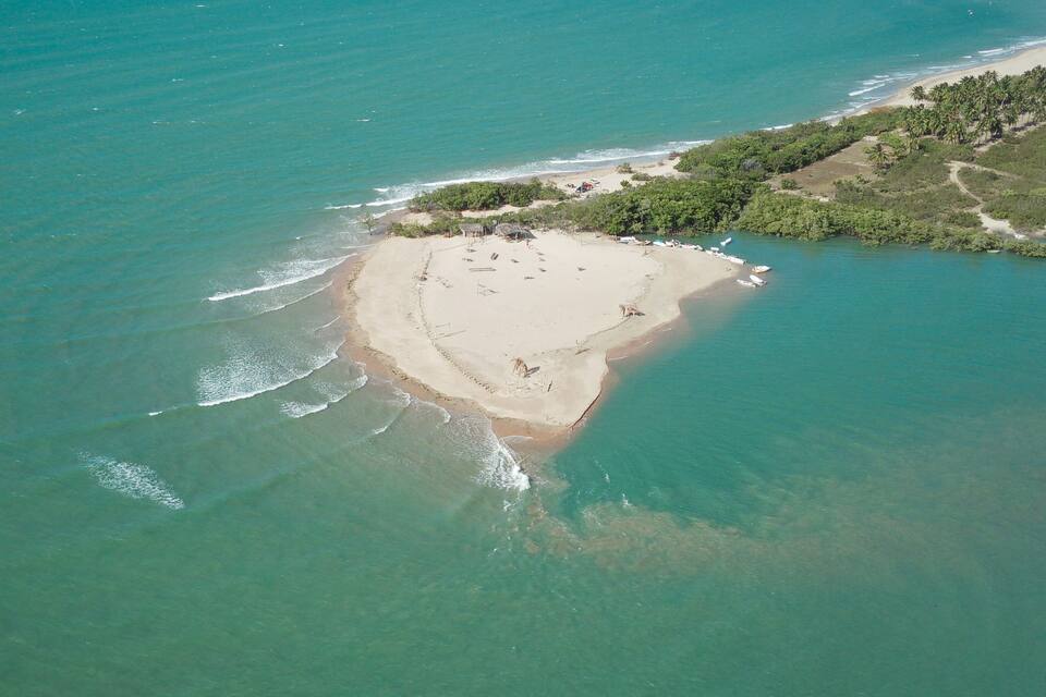 Vista aérea de una playa con aguas turquesas cristalinas, una orilla arenosa y un pequeño grupo de edificios rodeados de exuberante vegetación.