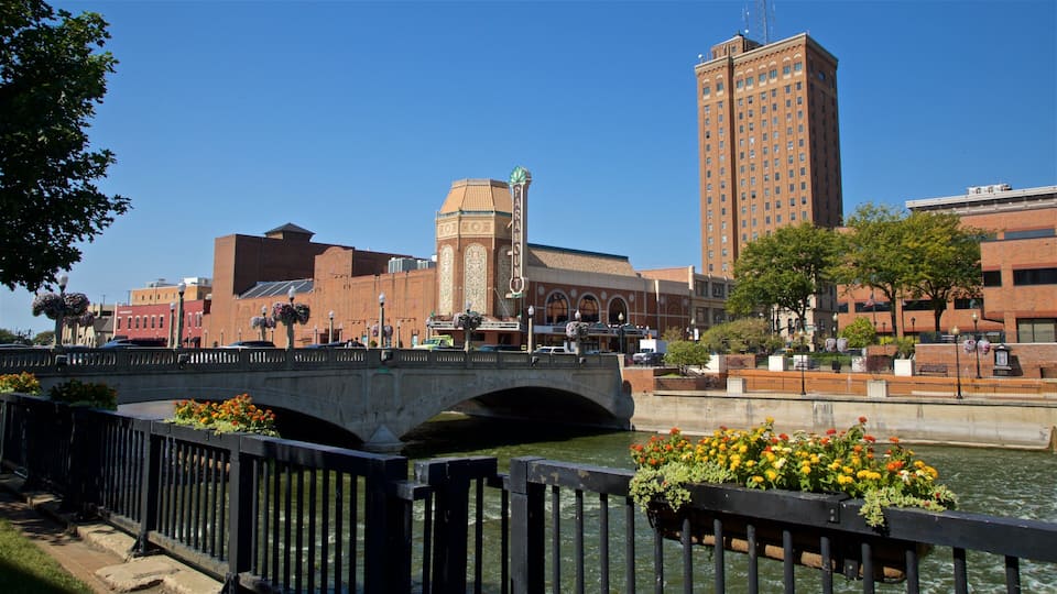 A bridge over a river with flower planters, a historic building with a clock tower, and a tall modern building in the background.