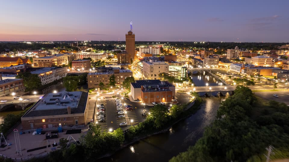 A cityscape at dusk with illuminated buildings, a river, and a prominent tower.