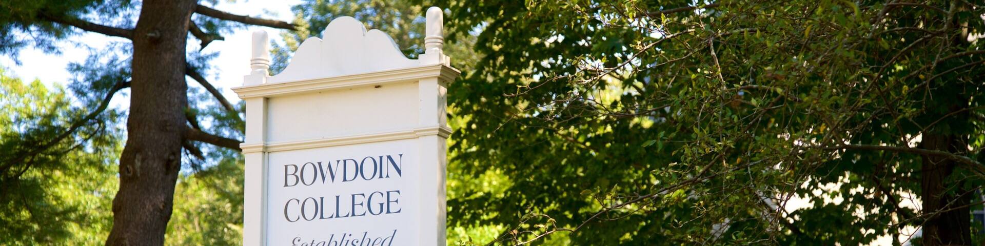 A sign for Bowdoin College with a tree and greenery in the background.