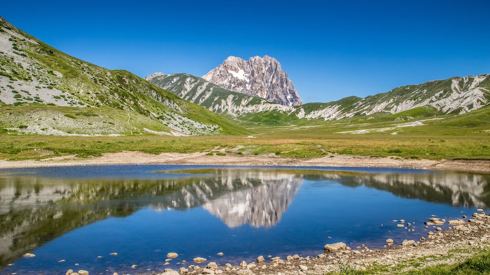 A mountain lake with clear reflections, surrounded by green hills and a prominent rocky peak.