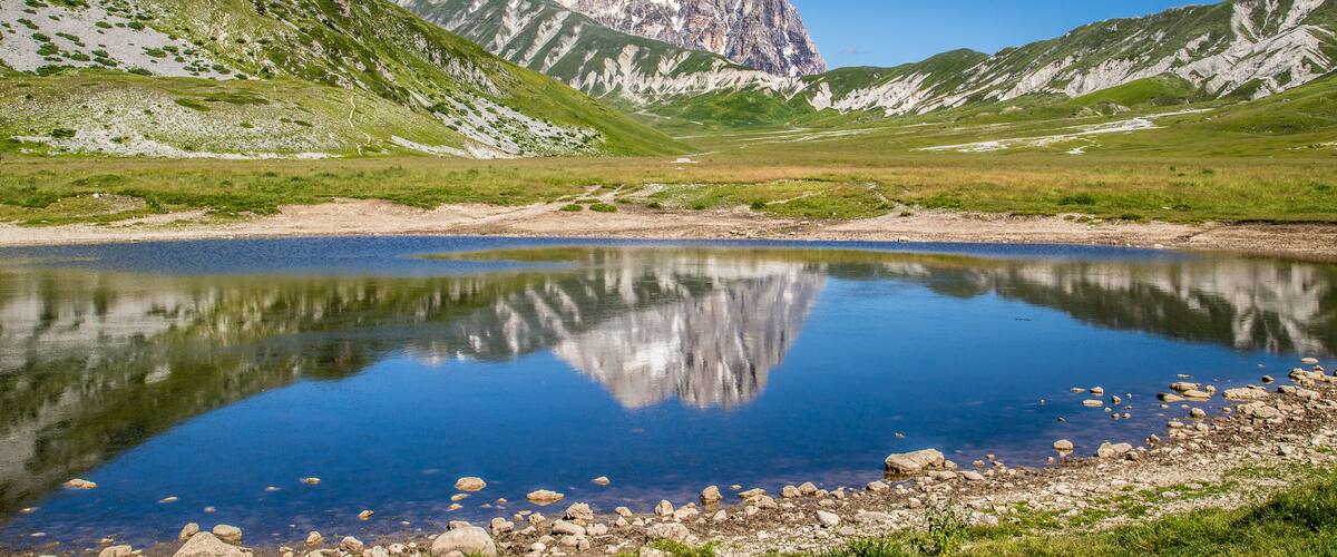 A mountain lake with clear reflections, surrounded by green hills and a prominent rocky peak.