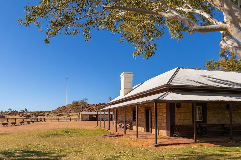 A historic stone building with a metal roof, surrounded by a grassy area with trees and a clear blue sky.