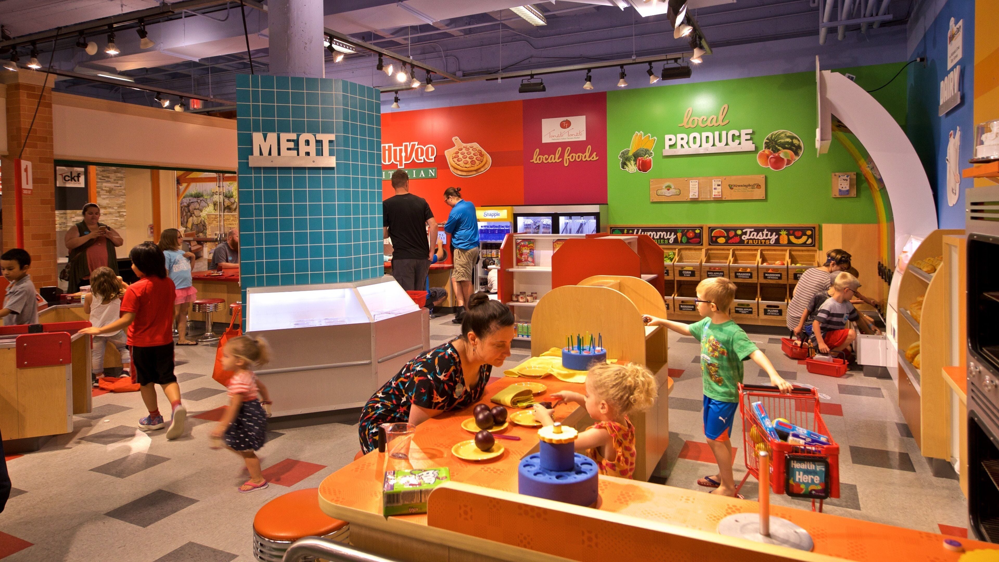 A colorful indoor play area with sections labeled 'MEAT' and 'LOCAL PRODUCE'.