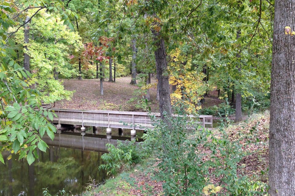 A wooden bridge over a calm river, surrounded by trees with autumn foliage.