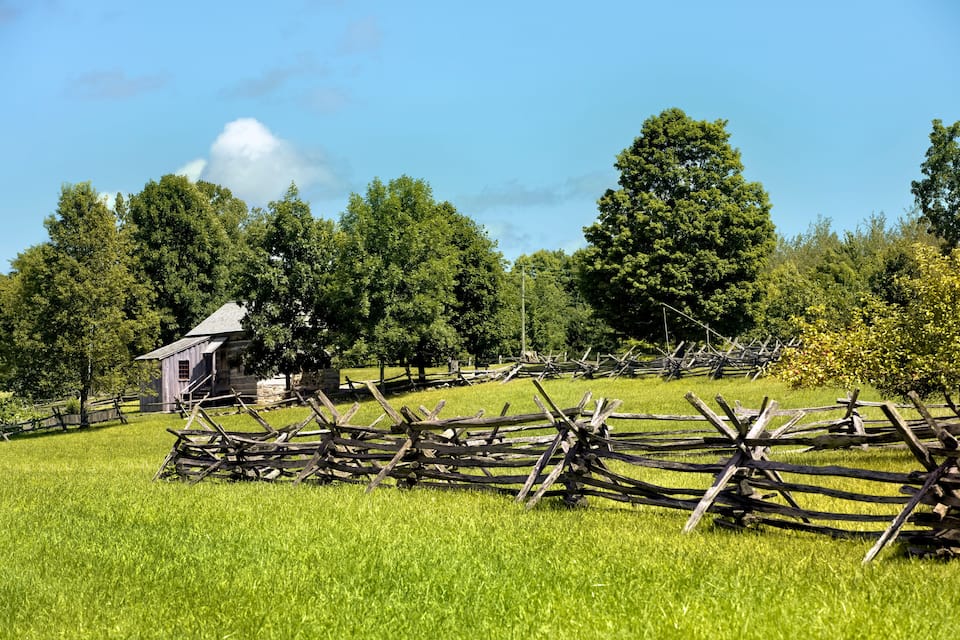 A rural landscape with a wooden fence, a small building, and lush green trees under a clear blue sky.