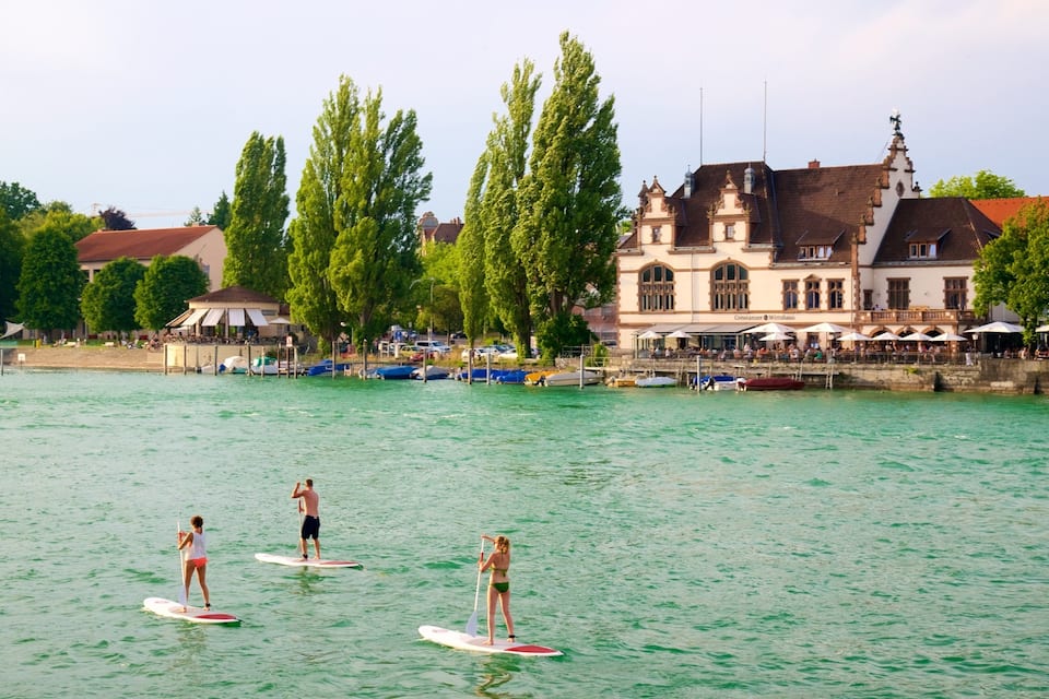 Tres persones fent paddleboard en un riu tranquil, amb un edifici històric i arbres al fons.