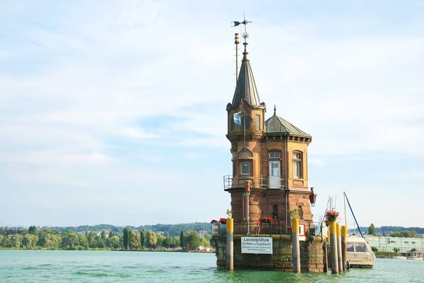 A historic tower on a floating platform in a lake, surrounded by greenery and a distant cityscape.