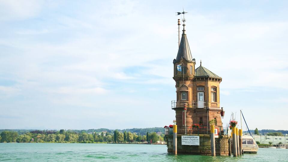 A historic tower on a floating platform in a lake, surrounded by greenery and a distant cityscape.