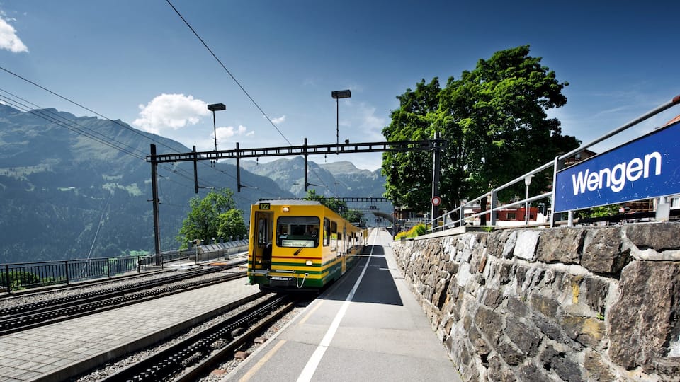 A yellow train at a station with a stone wall and a sign that reads "Wengen".