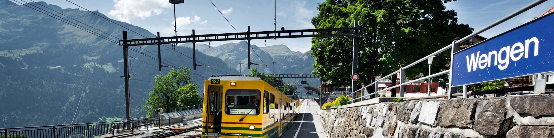 Un tren amarillo en una estación con un muro de piedra y un cartel que dice "Wengen".