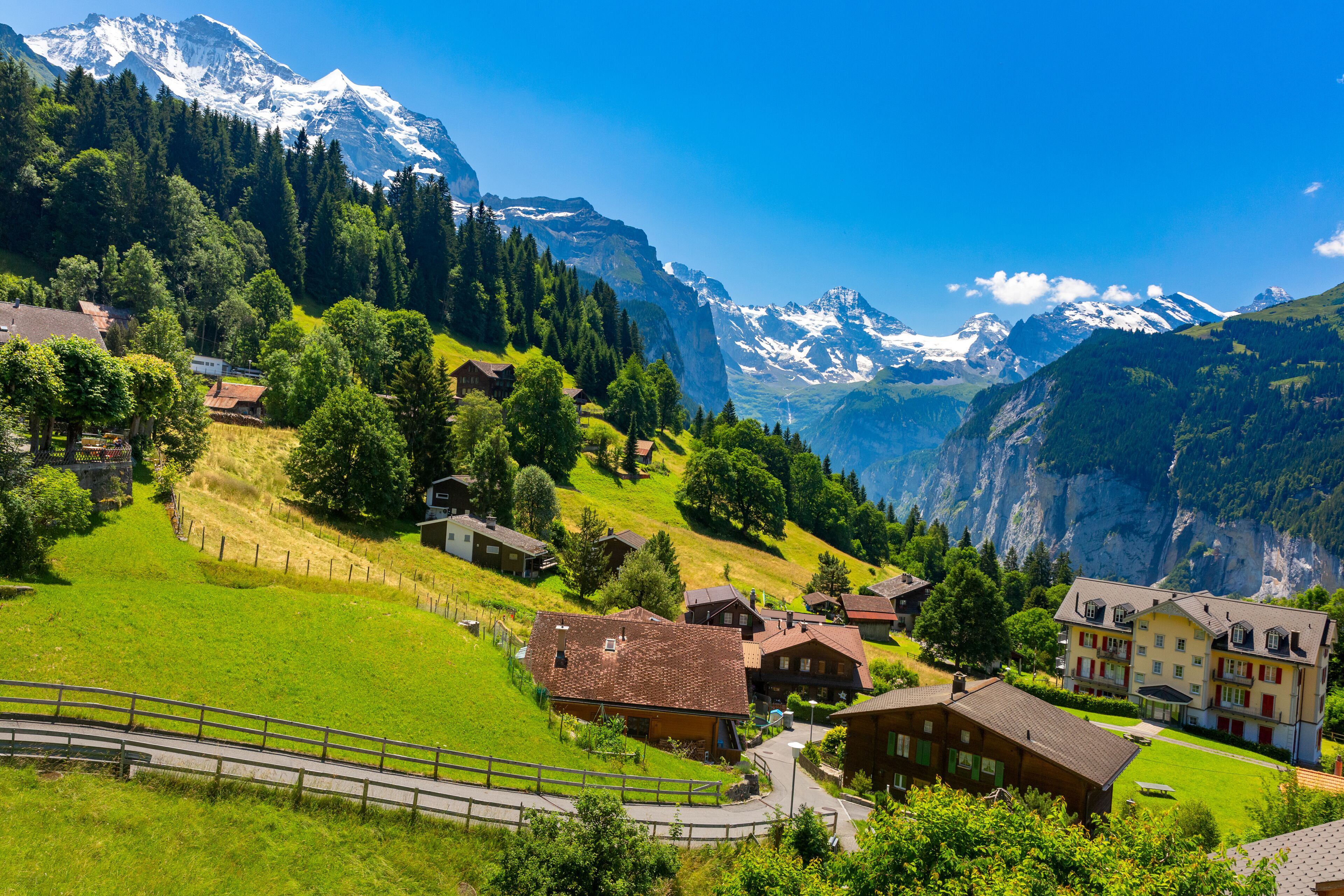 A mountainous village with houses, a winding road, and snow-capped peaks in the background.