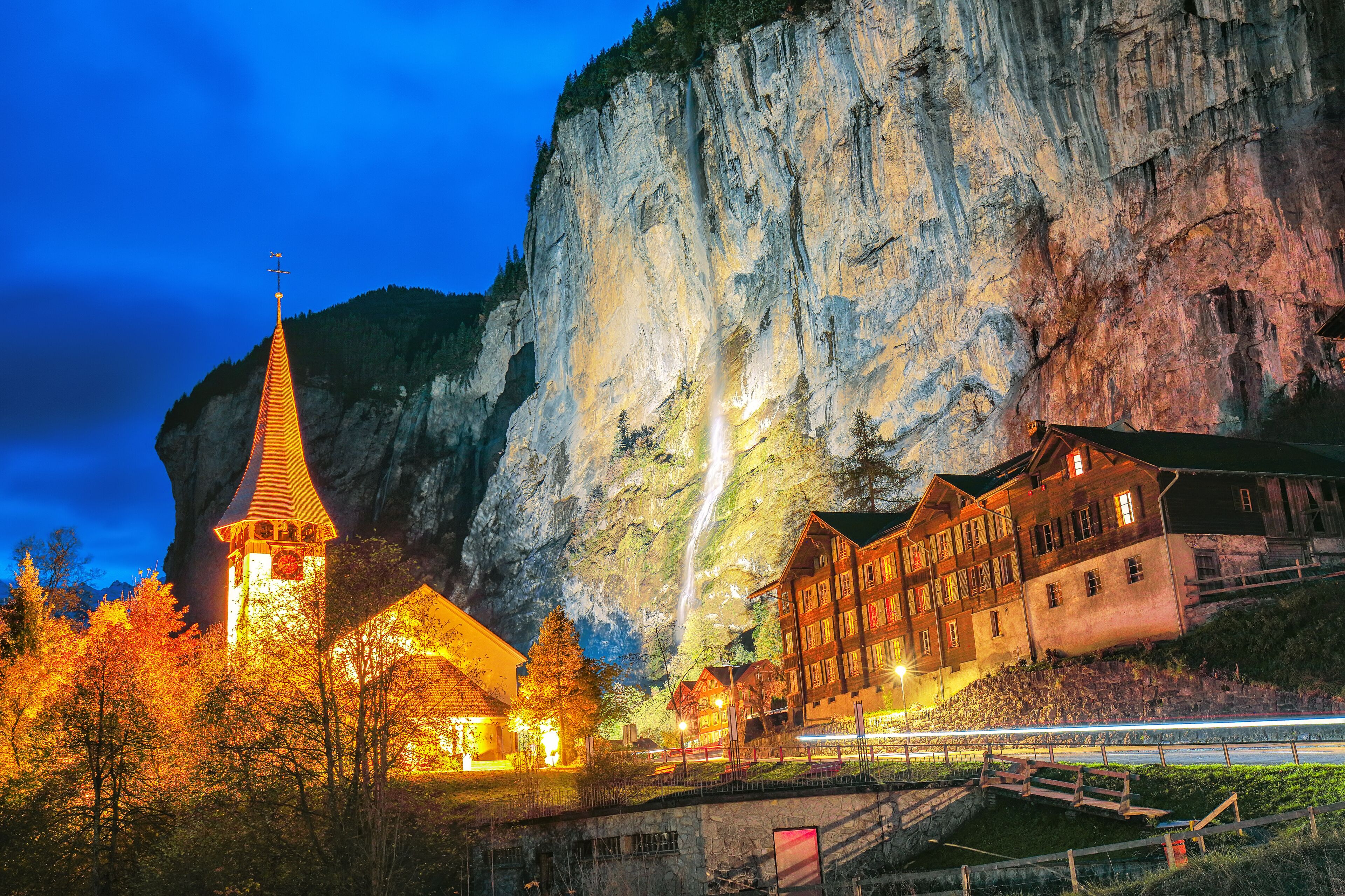 A church with a tall spire, a mountain backdrop, and a brightly lit building with multiple windows.