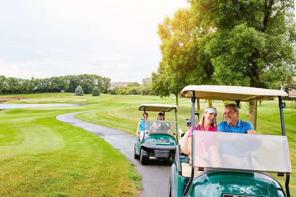 Un carrito de golf en un campo de golf con un camino verde, árboles y un cuerpo de agua al fondo.