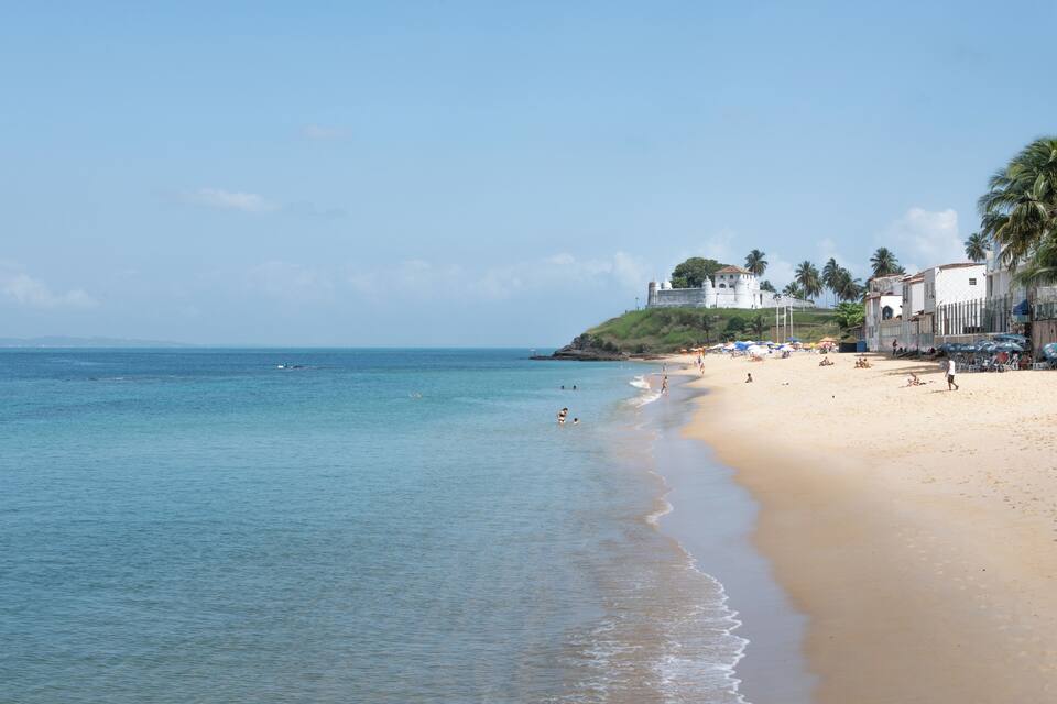 Une plage à l’eau d’un bleu cristallin, avec un rivage sablonneux et des bâtiments blancs bordés de palmiers.