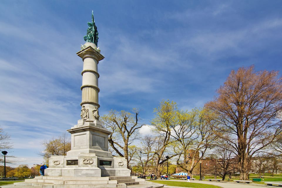 Ett högt monument med en staty på toppen, omgivet av träd och en klar, blå himmel.