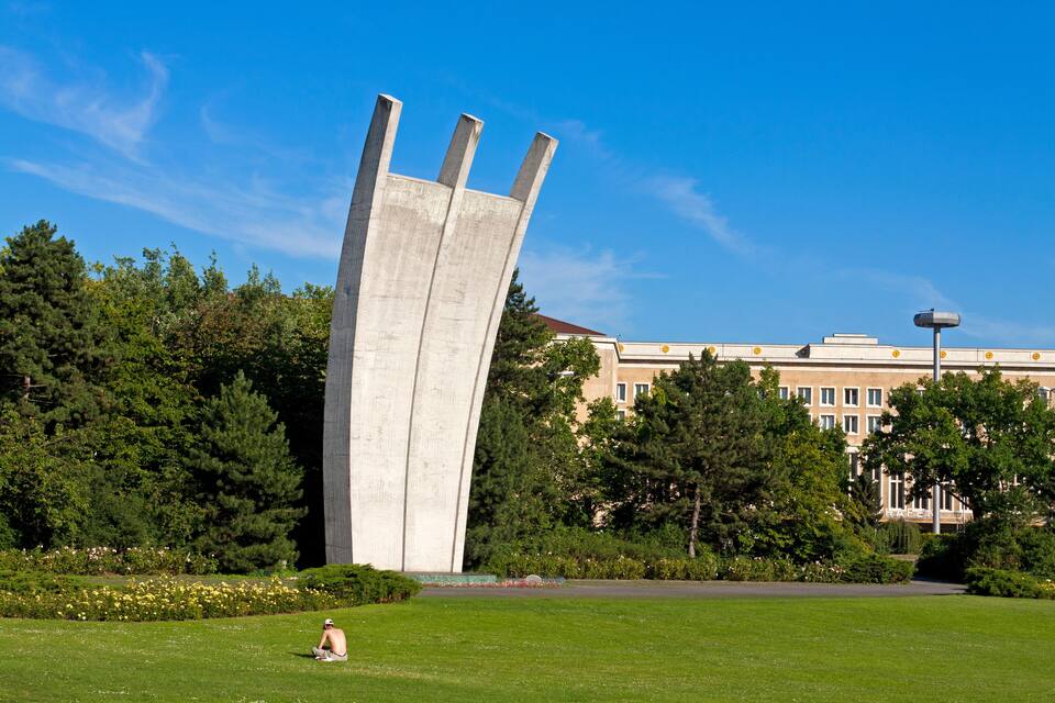 A tall concrete sculpture in a park with a building in the background.