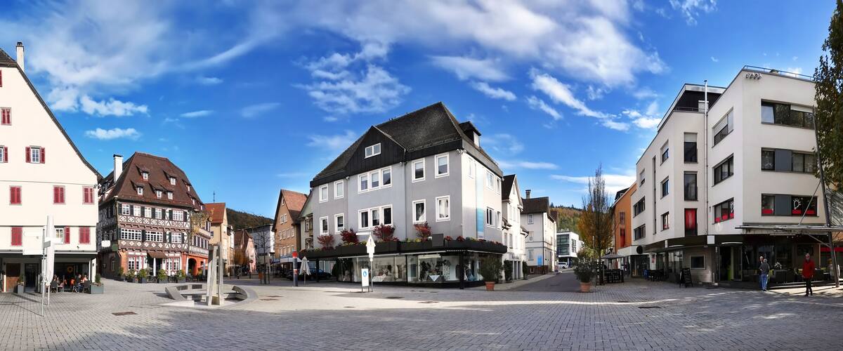 A cobblestone square with historic buildings, a central fountain, and a clear blue sky.