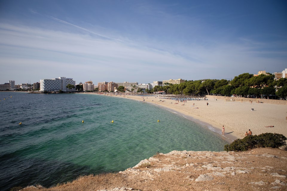 A coastal beach with clear turquoise waters, sandy shore, and buildings in the background.