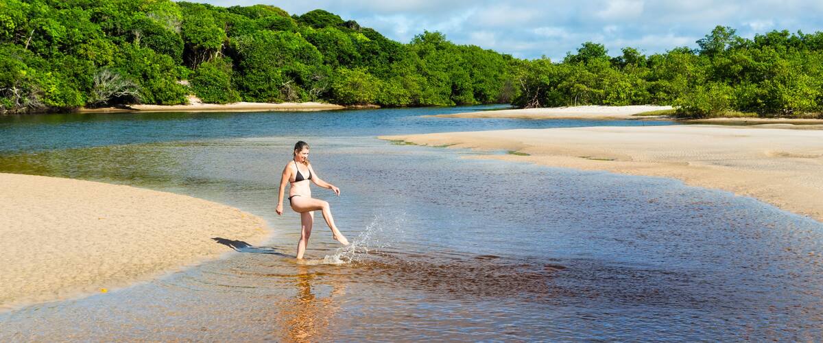 A woman in a bikini walking through shallow water near a sandy beach with dense green foliage in the background.