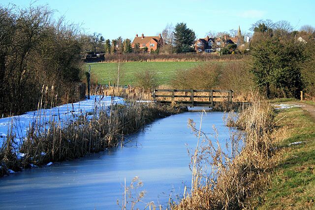 A frozen river with a wooden bridge, surrounded by trees and houses in the background.