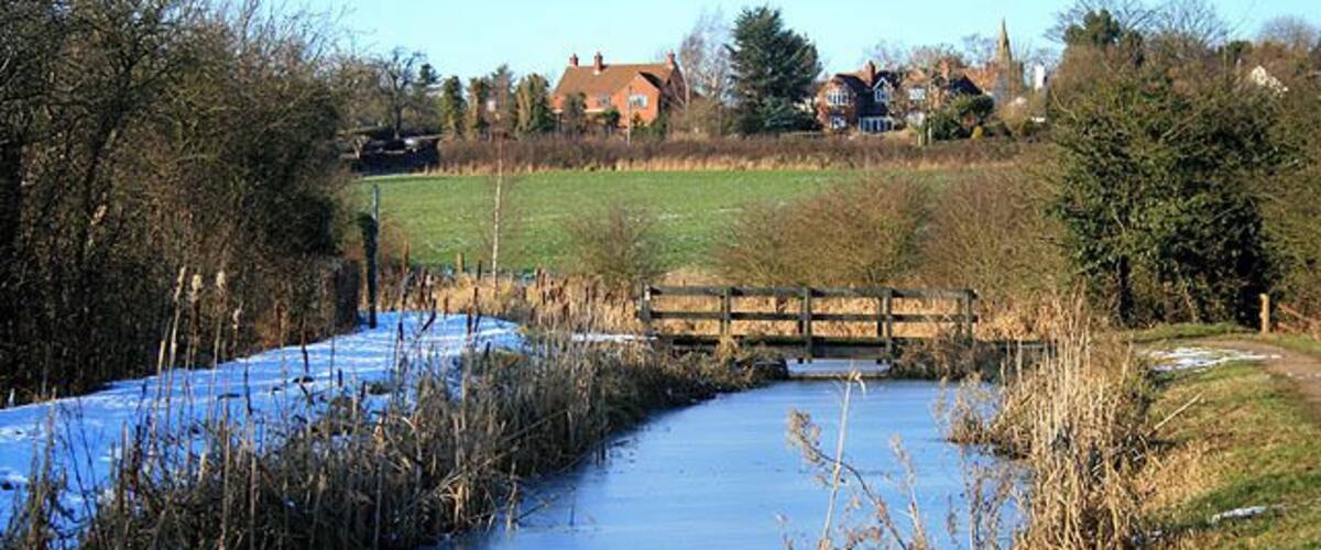 A frozen river with a wooden bridge, surrounded by trees and houses in the background.