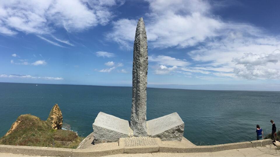 A tall, narrow stone monument with inscriptions, surrounded by a coastal landscape and the sea.