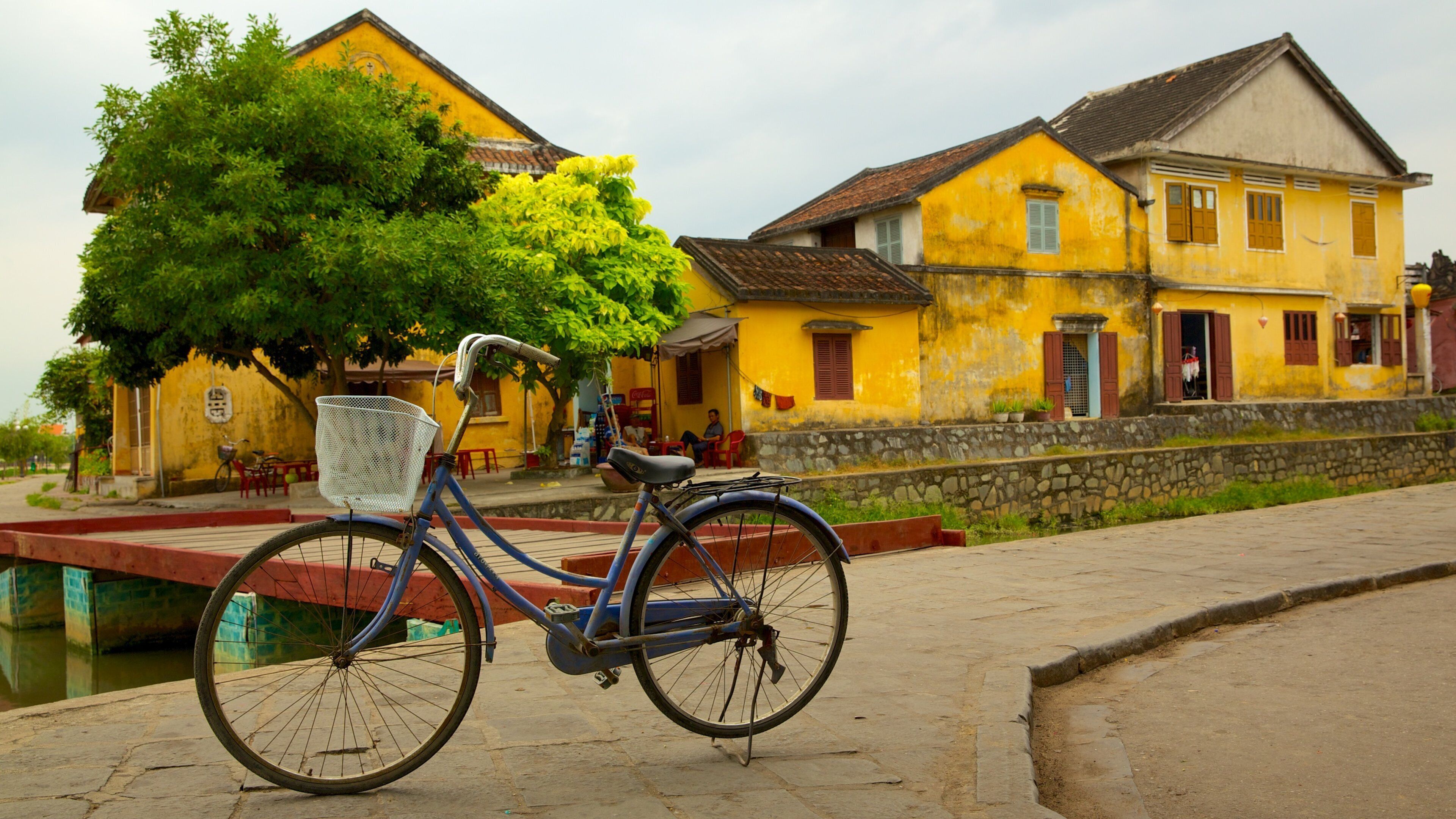 A blue bicycle parked on a stone path with a yellow building and a tree in the background.