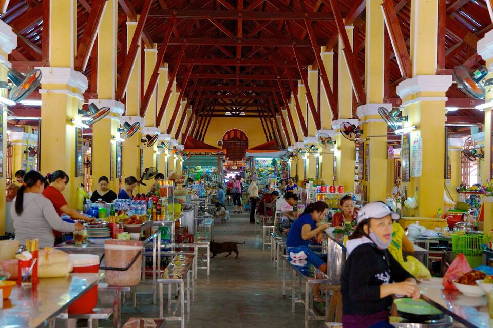 A bustling indoor market with yellow walls and red beams, featuring various stalls and people shopping.