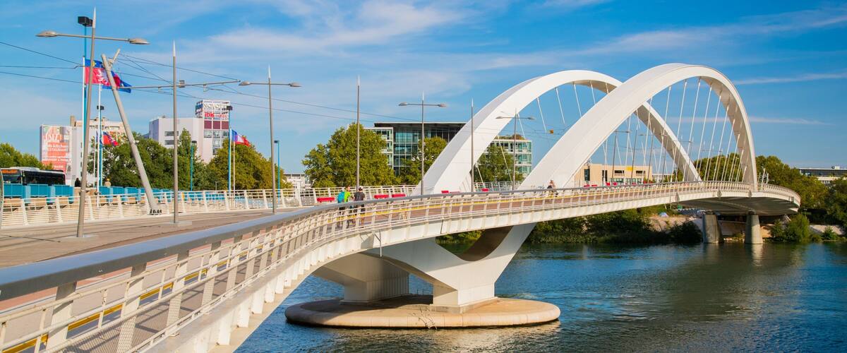 A modern bridge with two large arches spanning a river, surrounded by urban buildings and greenery.