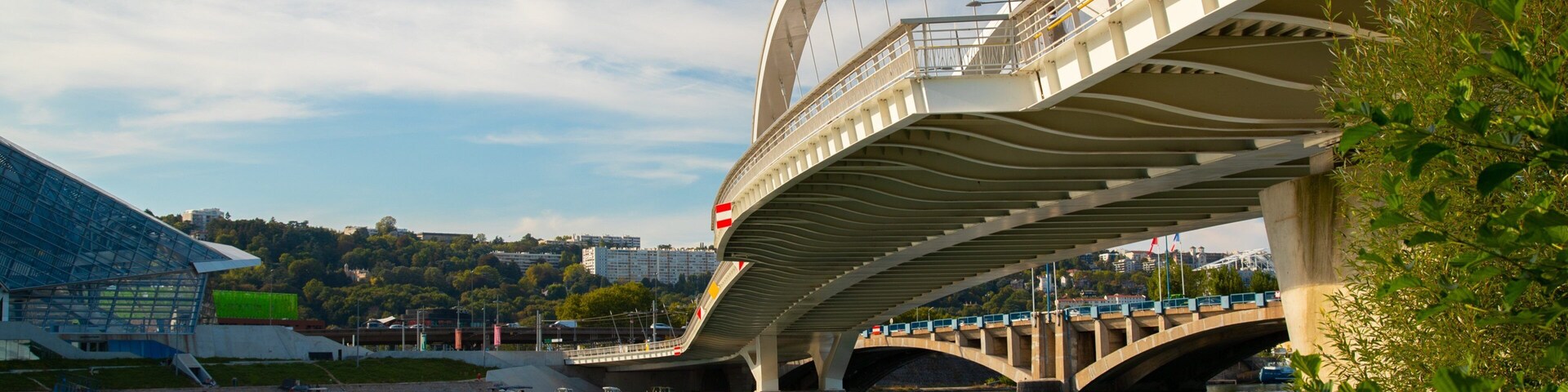 Uma ponte moderna com um design de arco único sobre um rio, com um estádio e edifícios ao fundo.
