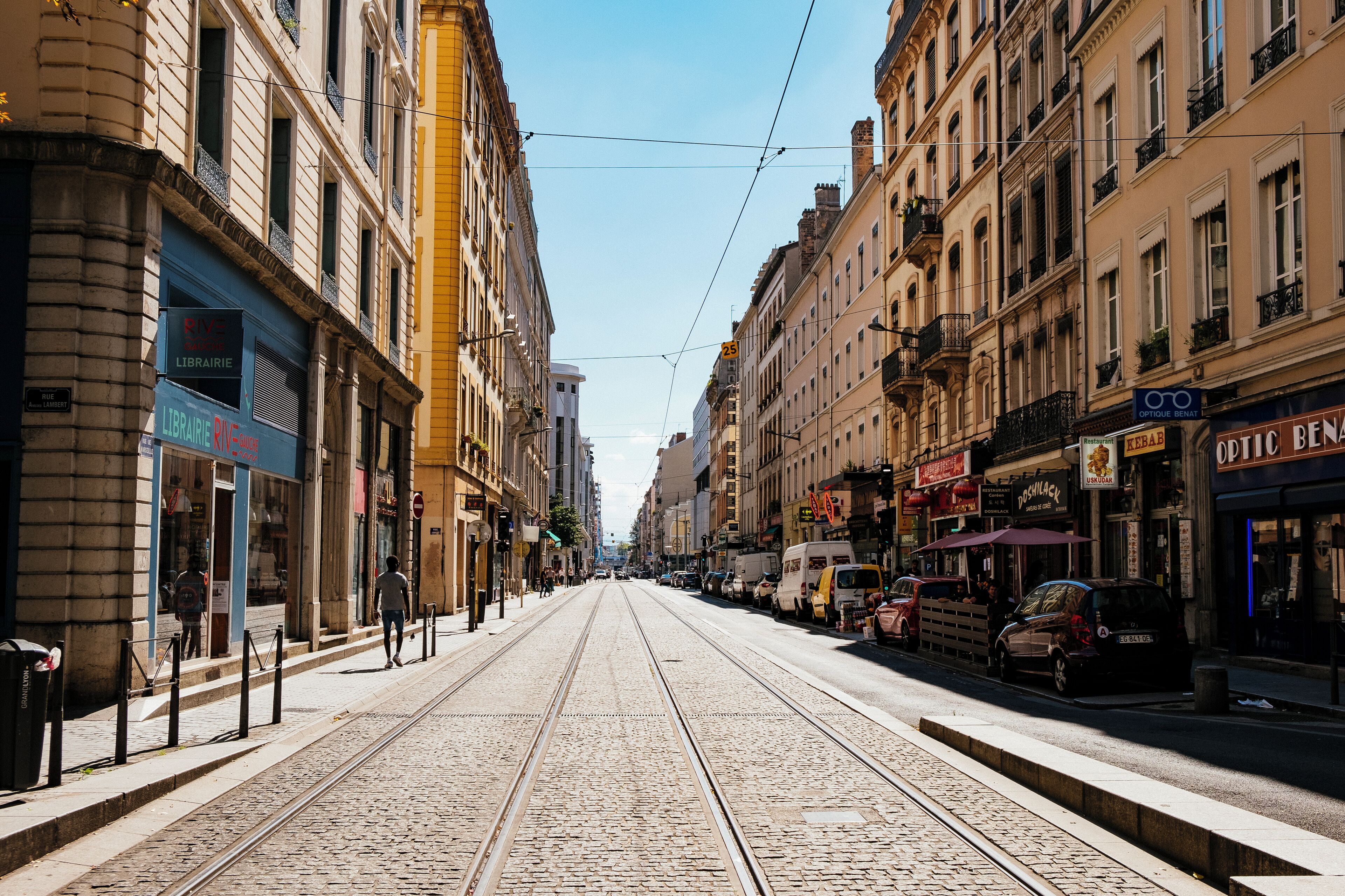 Uma rua de paralelepípedos com trilhos de bonde, edifícios com lojas e carros estacionados.