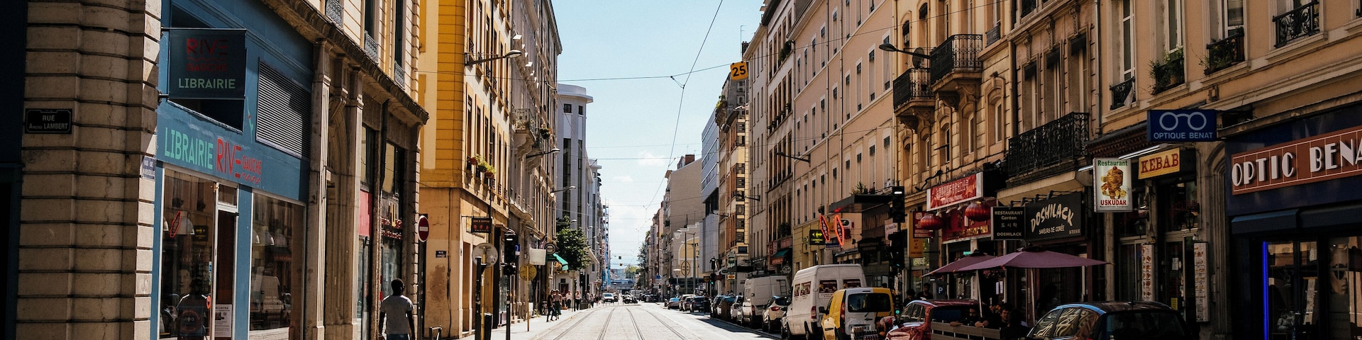 Uma rua de paralelepípedos com trilhos de bonde, edifícios com lojas e carros estacionados.